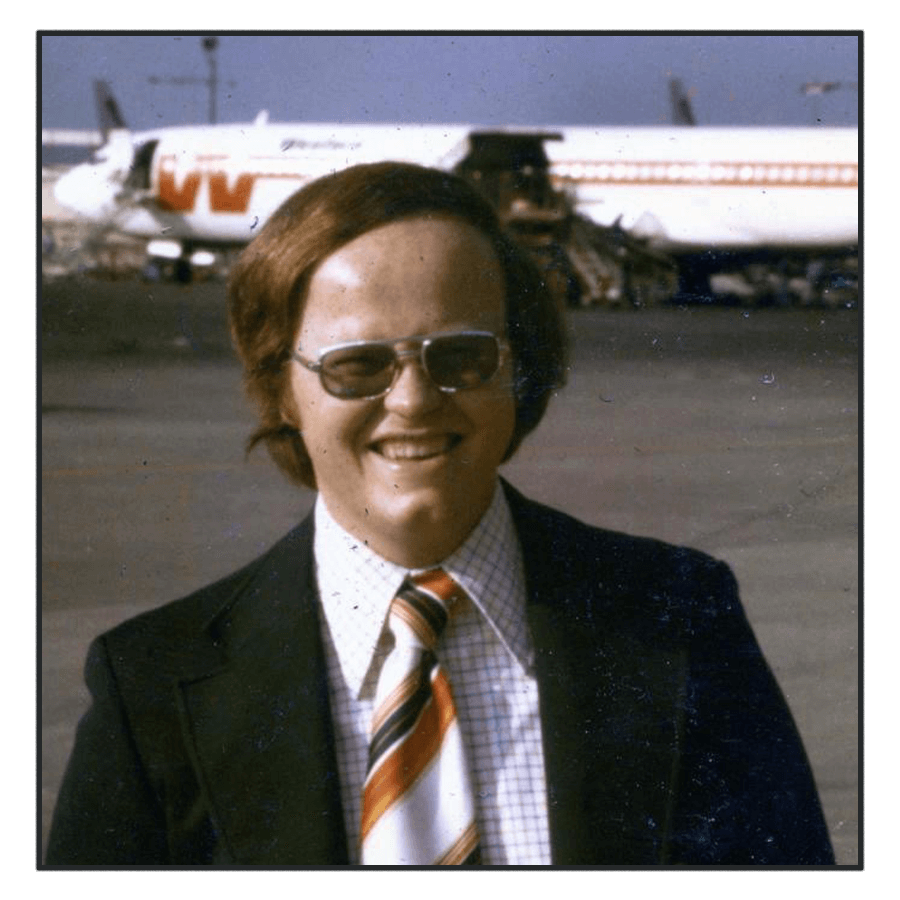 A man in a black suit smiling eagerly at the camera with an airplane in the background, representing the friendly and professional team at Cormorant Marketing, offering expert marketing services for aviation industry professionals.