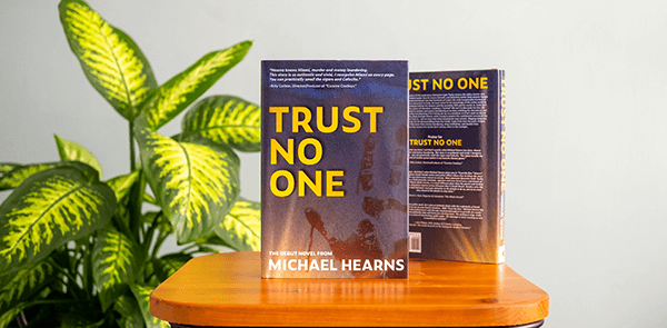 books Trust No One on a wooden platform with green plant in background