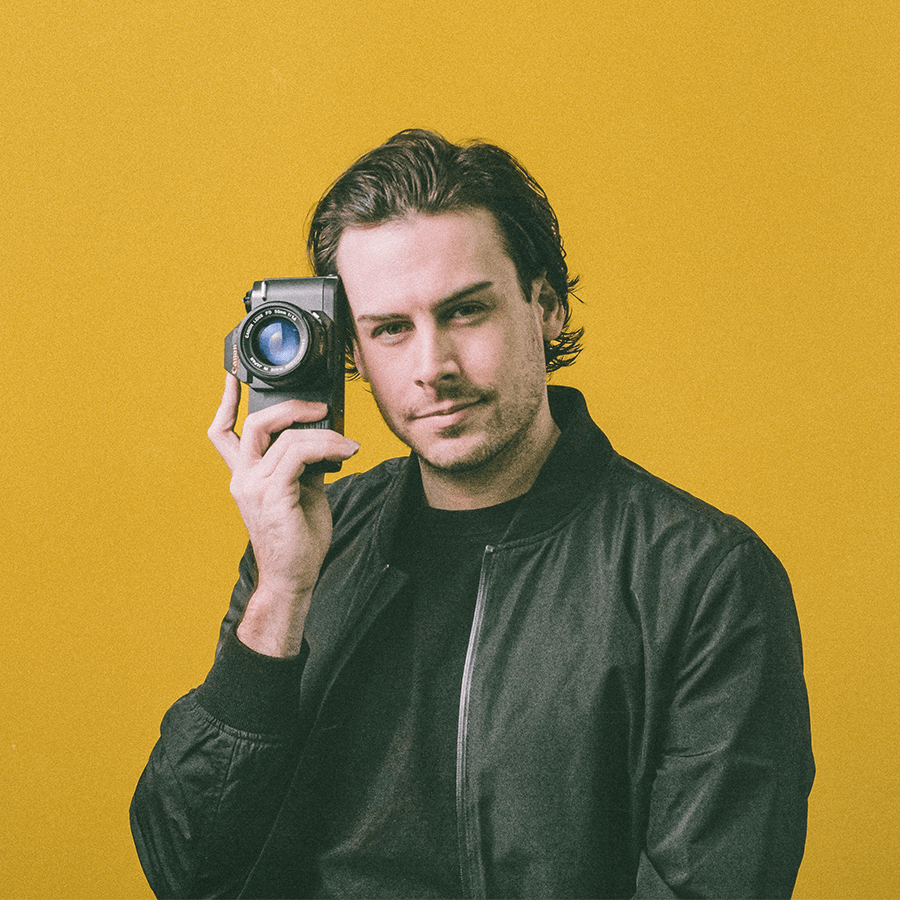 Dillon Hearns Portrait of the founder and CEO of Cormorant Marketing, a professional man with medium hair in a black jacket and tee, holding a camera thoughtfully against a striking gold background, symbolizing authority and expertise in marketing solutions for Jacksonville FL businesses.